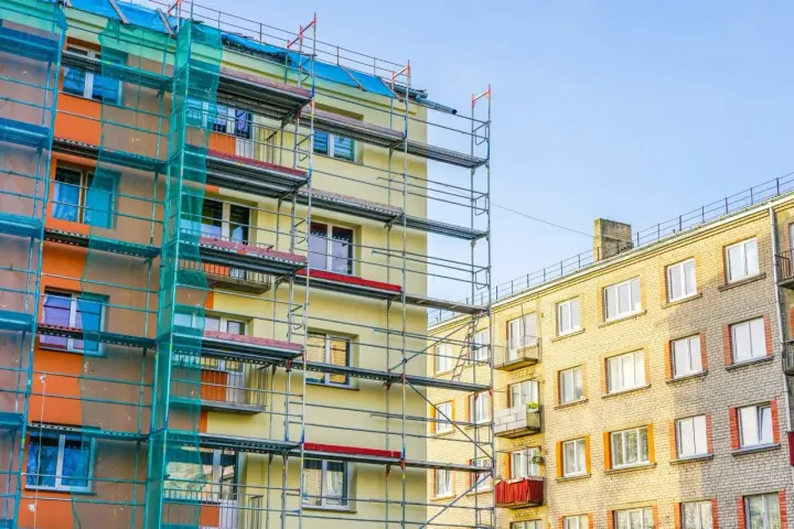 Budynek w remoncie, rusztowania i siatka ochronna. Czy balkon jest częścią mieszkania? Widać różne balkony, niektóre z czerwonymi markizami.