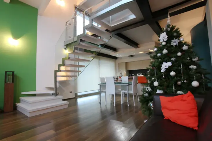 Un moderno soppalco casa con scala in vetro e legno, albero di Natale addobbato e un divano con cuscino arancione.