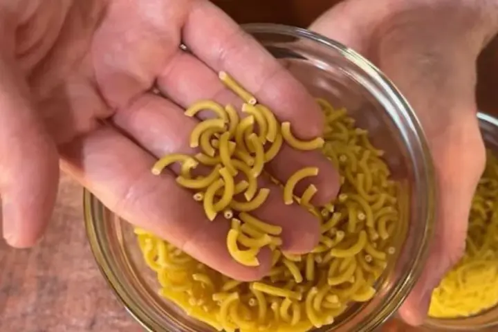 Mano sosteniendo fideos cortos y curvos sobre un bol de fideos similares.