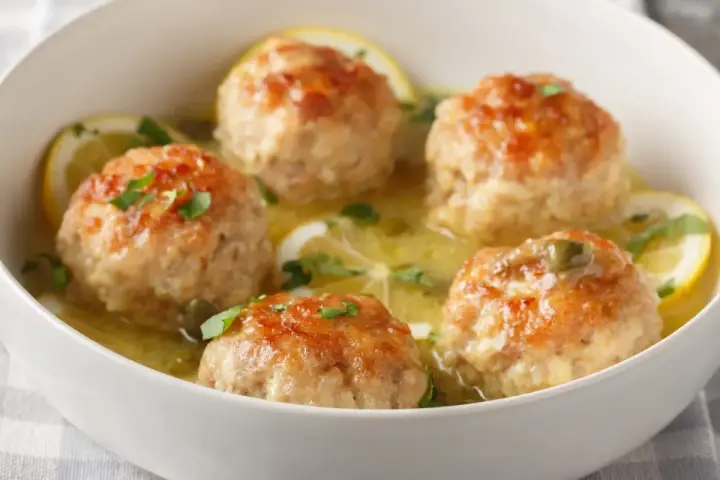 Polpette al limone dorate e succulente, servite in una ciotola bianca con fette di limone e prezzemolo tritato.