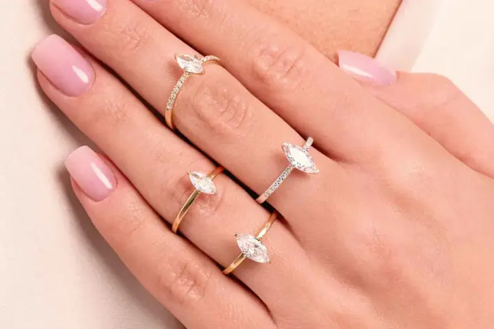 Dłonie z różowym manicure ozdobione są trzema złotymi pierścionkami z białymi kamieniami. Jeden z nich to pierścionek kwadratowy.