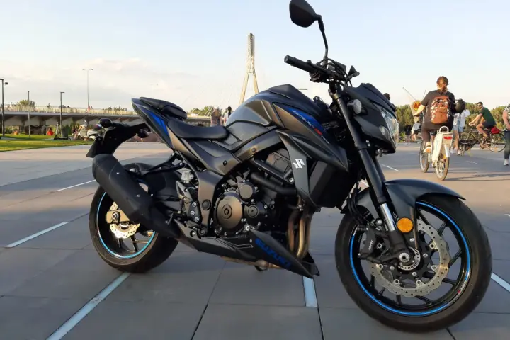 Czarny motocykl Suzuki GSX-S 750 z niebieskimi akcentami na felgach i owiewkach, zaparkowany na placu z widocznym w tle mostem i ludźmi.