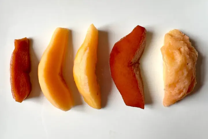 Cinq morceaux de fruit coing cuit, allant du rouge profond à l'orange clair, disposés en ligne sur fond blanc.