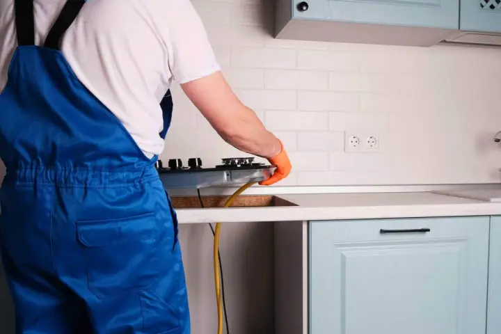 Instalator w niebieskim kombinezonie podłącza kuchenkę gazową w kuchni.