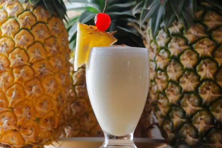 Pina Colada na mleczku kokosowym czy na śmietance? Orzeźwiający drink z ananasem i wisienką, otoczony dojrzałymi owocami.