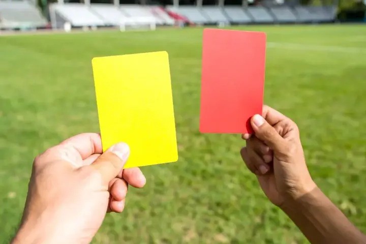 Sędzia pokazuje żółtą kartkę, która oznacza ostrzeżenie w piłce nożnej. Obok czerwona kartka.