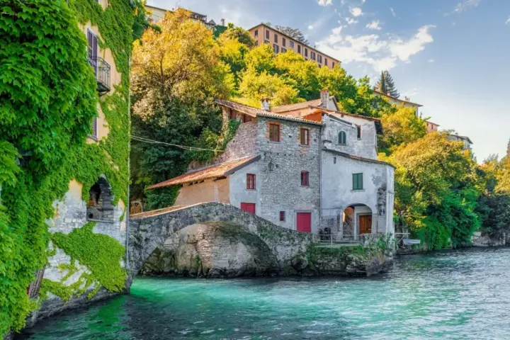 Antica casa in pietra con edera su un lago turchese, un ponte ad arco e colline verdi. Un luogo incantevole per i cammini in Lombardia.