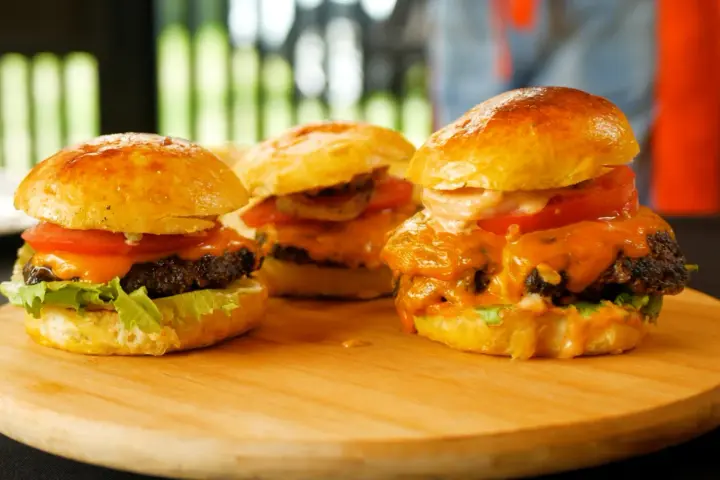 Trzy soczyste burgery z roztopionym serem, pomidorem i sałatą na desce.