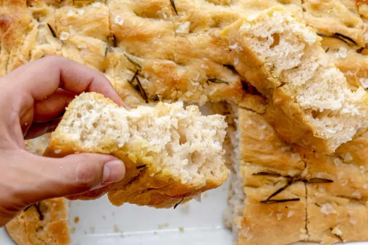 Mano rompiendo un trozo de focaccia casera con romero y sal gruesa. ¡Una delicia de esta focaccia receta!
