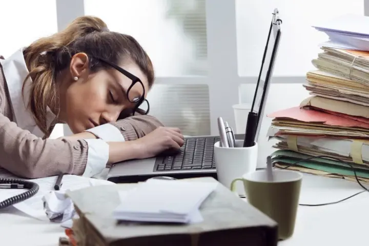 Zmęczona kobieta śpi na laptopie, otoczona stertami dokumentów. To obrazek pokazujący skutki związku z pracoholikiem.