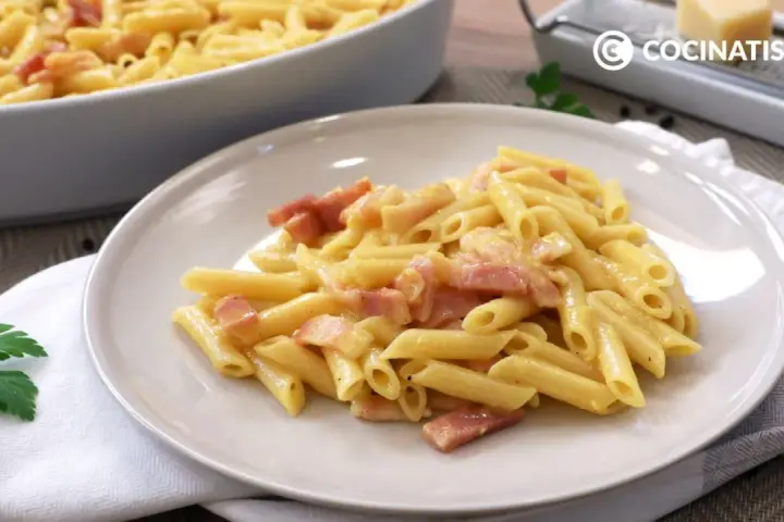 Plato de macarrones a la carbonara cremosos con trozos de panceta. En el fondo, un bol con más pasta y un rallador de queso.