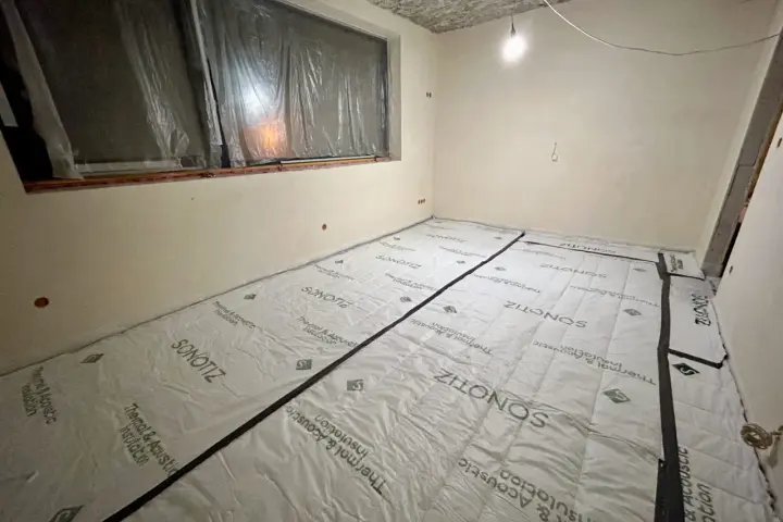W trakcie prac remontowych w bloku, podłoga pokryta jest materiałem do wyciszenia. Widać okno zabezpieczone folią i sufit z widocznymi instalacjami.