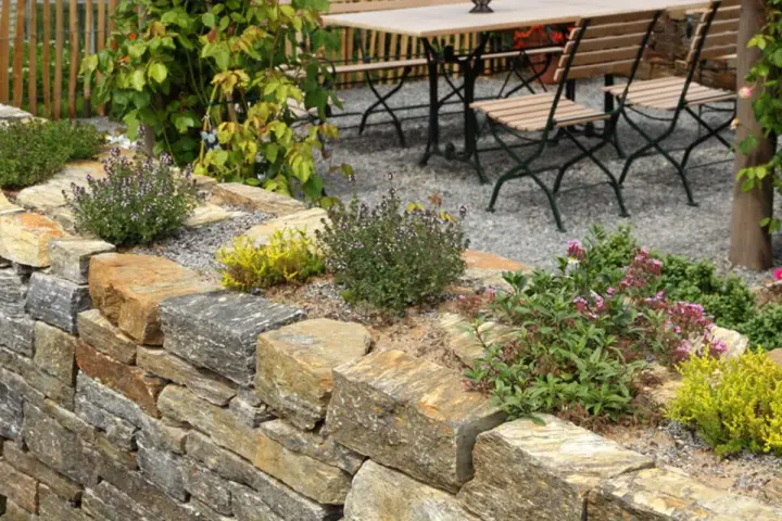 Natürliche Steinmauer mit Kräutern und Blumen, die schöne Ideen für den Garten bietet. Eine Sitzgruppe lädt zum Verweilen ein.
