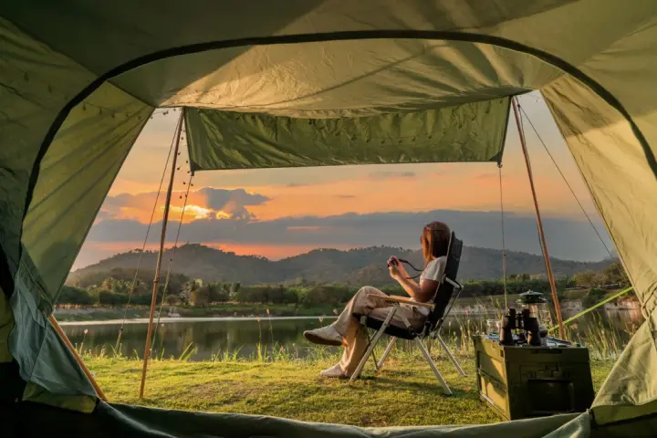 A woman enjoys solo camping by a lake at sunset, holding a camera. The tent offers a cozy view of the serene landscape.
