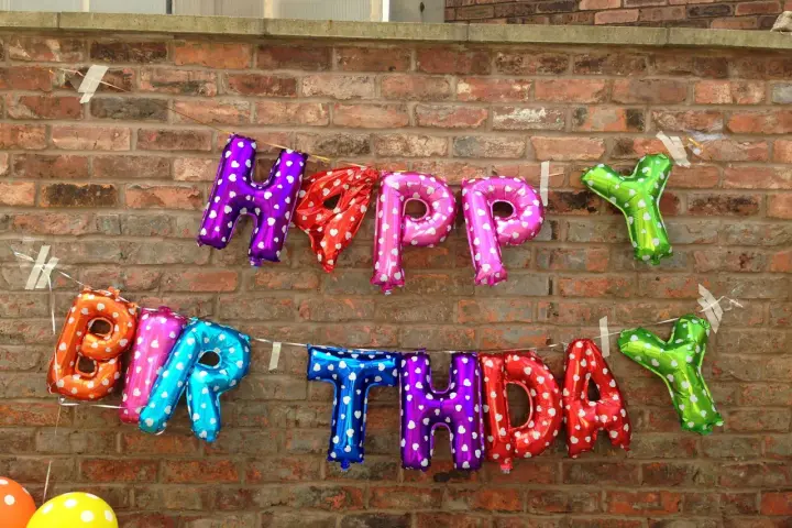 Luftballons in Buchstabenform bilden "HAPPY BIRTHDAY" an einer Ziegelwand.