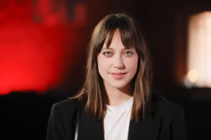 Maria Sobocińska z niebieskimi oczami i grzywką, ubrana w czarną marynarkę i białą koszulkę, uśmiecha się lekko na tle czerwonego światła.