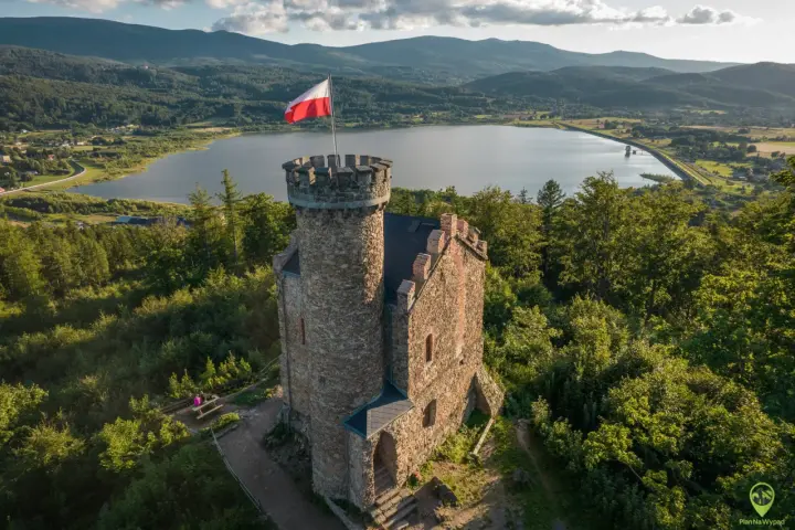 Kamienny zamek księcia Henryka z polską flagą na wieży, otoczony lasem, z widokiem na jezioro i góry. Punkt widokowy zaprasza do podziwiania krajobrazu.