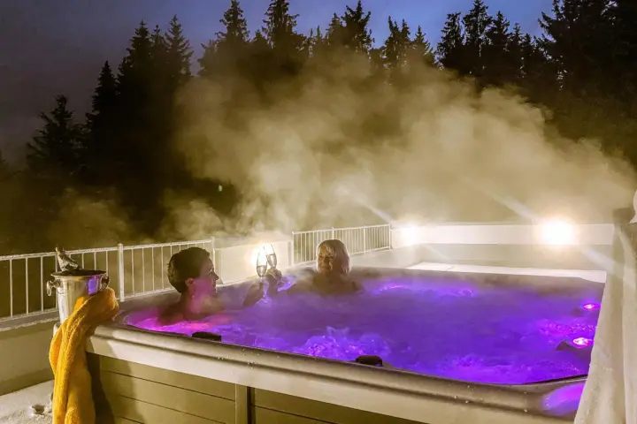 Zwei Frauen stoßen mit Champagner in einem beleuchteten Whirlpool an, umgeben von Dampf und Bäumen. Ein perfektes Wochenende mit Gott.