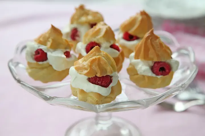 Små petit choux fyllda med grädde och hallon på ett glasfat.