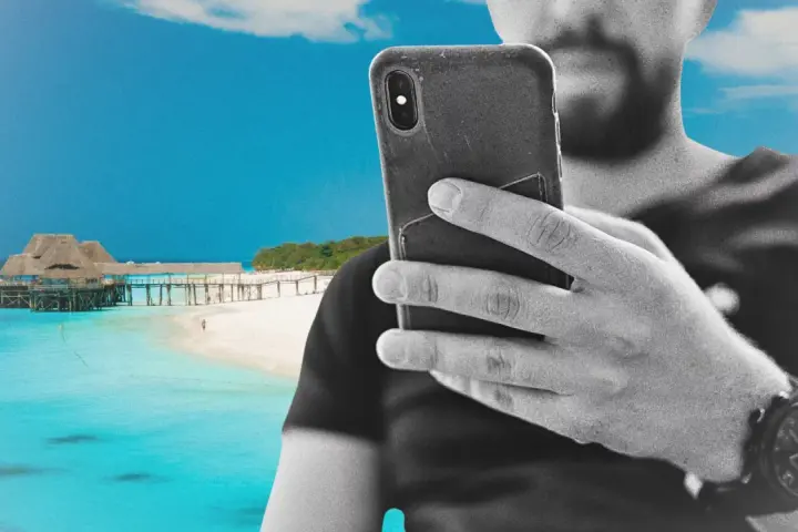 Mężczyzna z telefonem na plaży na Zanzibarze, sprawdzający zanzibar internet. W tle turkusowa woda i drewniany pomost.