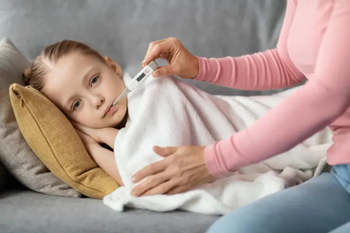 Dziecko leży pod kocem, a mama mierzy mu temperaturę. Wysoka gorączka u dziecka bez innych objawów martwi.