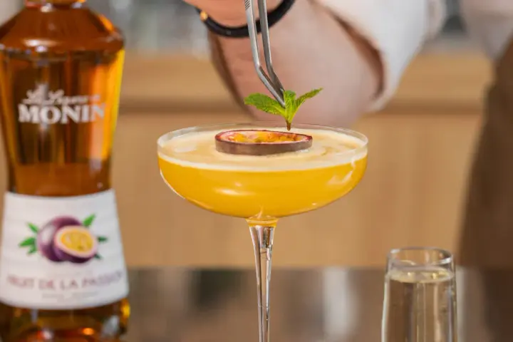 Barman dekoruje drink likierem z marakui, dodając listek mięty. Obok stoi butelka Monin.