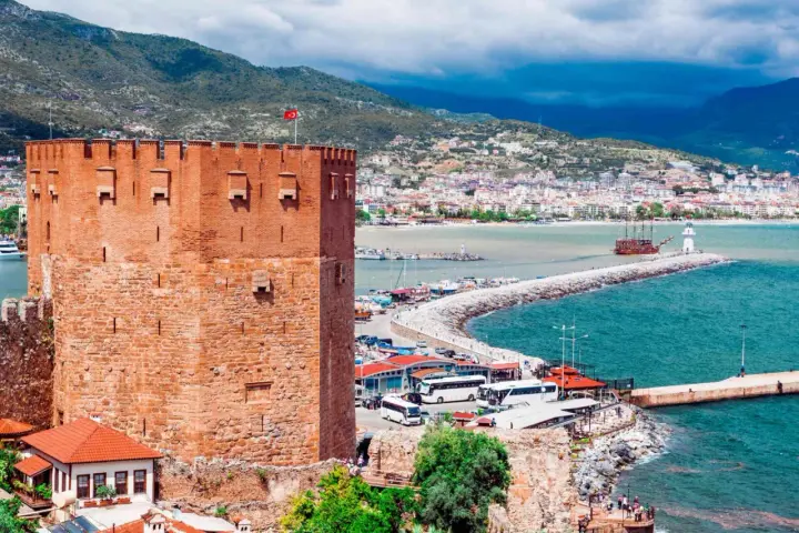 Alanya zwiedzanie na własną rękę: potężna czerwona wieża zamku, port z łodziami, latarnia morska i miasto w tle.