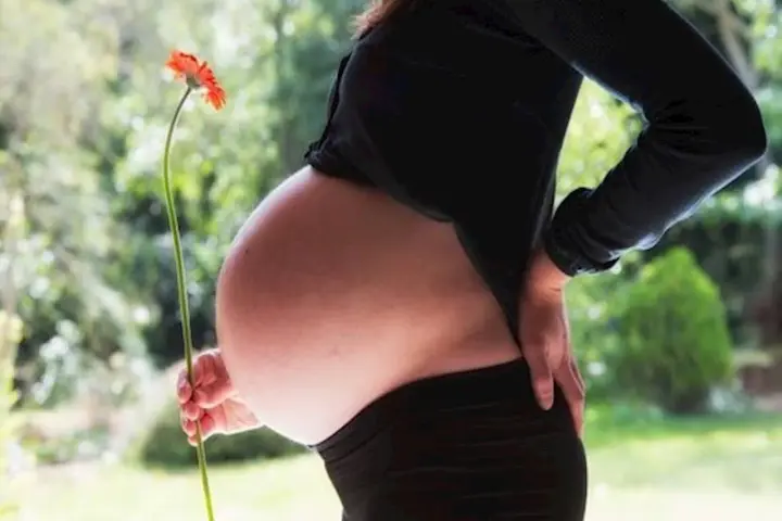 Brzuszek w 3 miesiącu ciąży, delikatnie podtrzymywany dłonią, z kwiatkiem w tle.