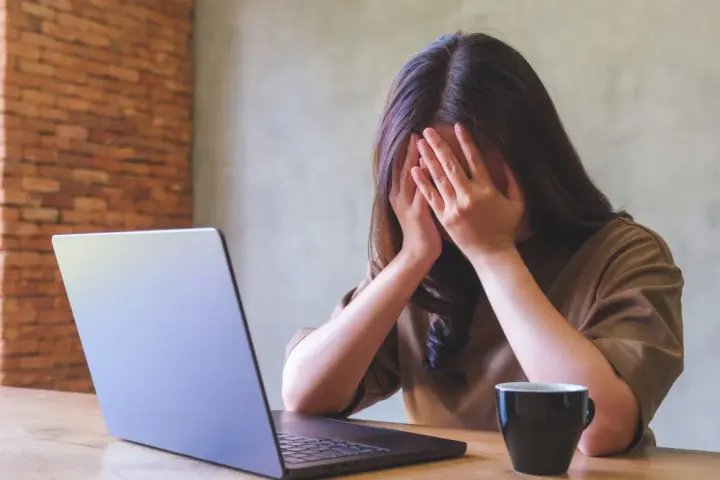 Kobieta z dłońmi na twarzy, przy laptopie, z kubkiem kawy. Objawy wypalenia zawodowego, czas na L4.