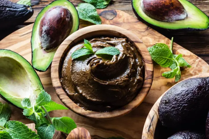 Crema di avocado vellutata in ciotola di legno, circondata da avocado freschi e foglie di menta.