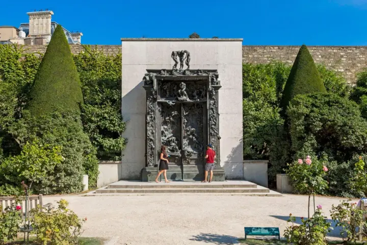 La Porte de l'Enfer, une œuvre monumentale de Rodin, se dresse majestueusement, encadrée par des arbres taillés et un mur de pierre. Deux visiteurs contemplent cette sculpture complexe.