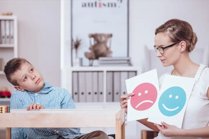 Bambino con autismo durante un test, mentre un terapista mostra faccine felici e tristi.