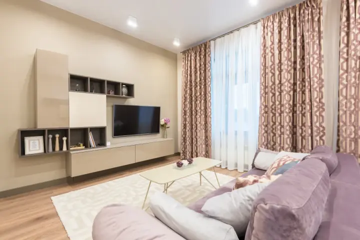 Przytulny salon z fioletową kanapą, beżowy kolor ścian tworzy ciepłą atmosferę. W tle telewizor i zasłony.