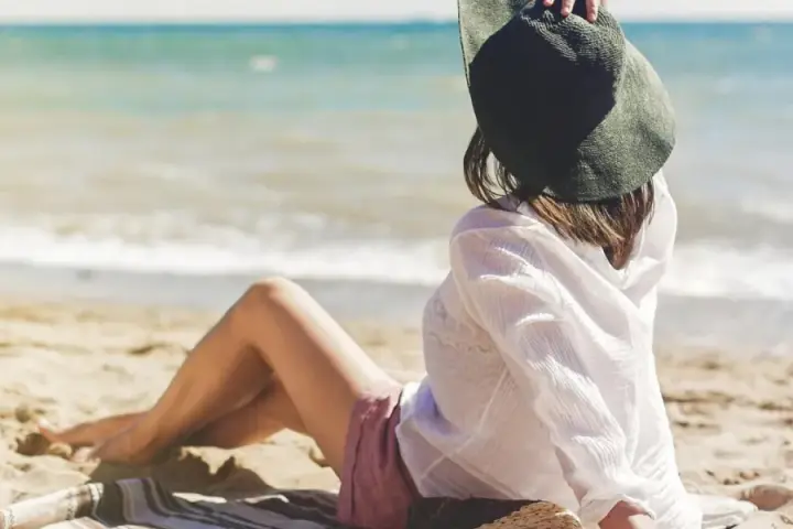 Kobieta w kapeluszu siedzi na plaży, w tle widać spokojne jezioro. Idealne miejsce na ustronny domek nad jeziorem.