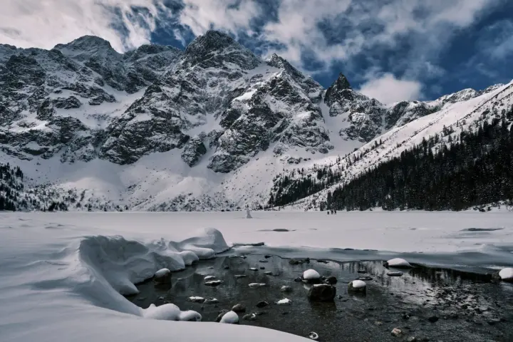 Zamarznięte Morskie Oko, otoczone ośnieżonymi szczytami Tatr. W oddali widać ludzi spacerujących po lodzie, a na pierwszym planie płynie strumyk.