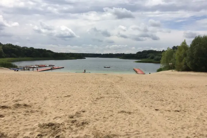 Piaskowa plaża nad jeziorem, z pomostami i łódkami. W oddali las, chmury na niebie. Idealne miejsce na wypoczynek nad wodą.