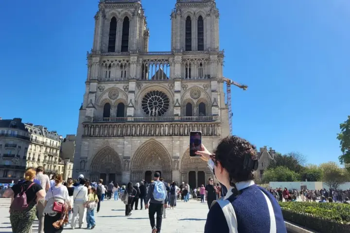 Una persona toma una foto de la fachada de Notre Dame. Mucha gente pasea por la plaza, disfrutando de su visita a Notre Dame.