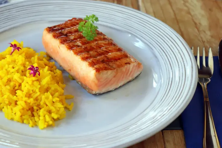Un delicioso salmón a la plancha con marcas de parrilla, servido con arroz amarillo y decorado con perejil y flores moradas.