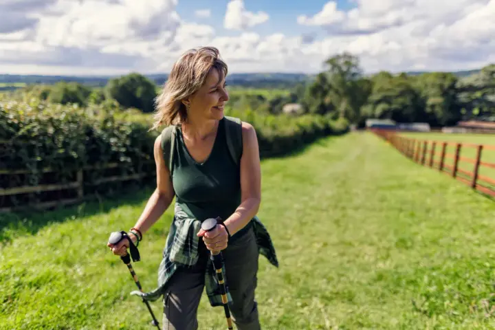 Donna sorridente con bastoncini da nordic walking, che lavora i muscoli coinvolti in un paesaggio verdeggiante.