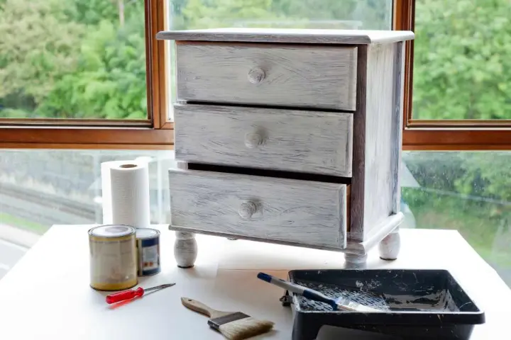 Mueble de tres cajones con acabado desgastado, listo para pintar muebles oscuros. Pinceles, rodillo y latas de pintura a su lado.