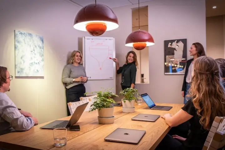 Ett team diskuterar strategier vid ett konferensbord med flera laptops. En kvinna pekar på en trattmodell på en whiteboard. Ups datorn är en del av deras effektiva arbetsflöde.