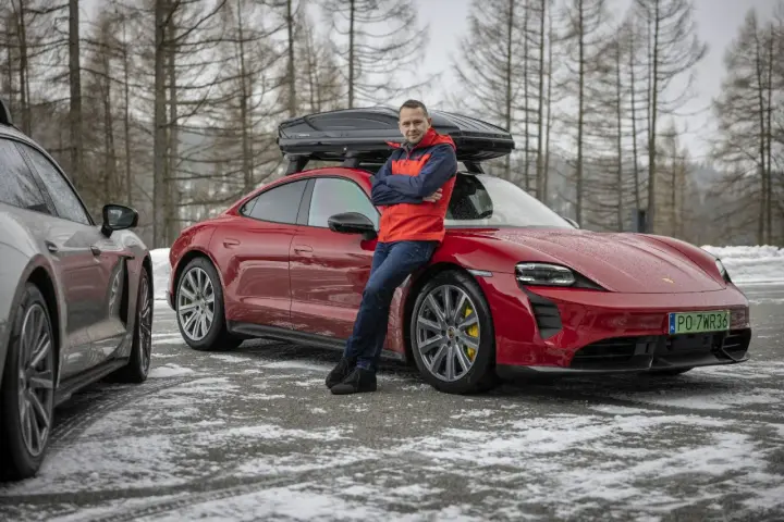 Mężczyzna w czerwonej kurtce opiera się o czerwonego Porsche Taycan z boxem dachowym.