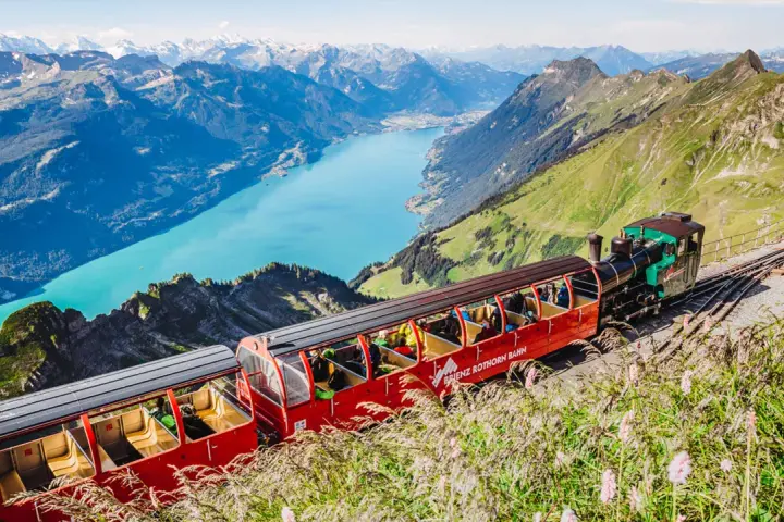 The Brienzer Rothorn train ascends a steep mountain track, offering passengers breathtaking views of a turquoise lake and snow-capped peaks.