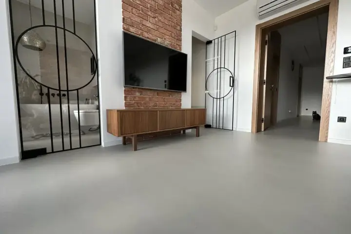 Nowoczesne wnętrze z betonową podłogą, co to jest mikrocement, która nadaje mu surowego charakteru. W tle widać łazienkę i salon.
