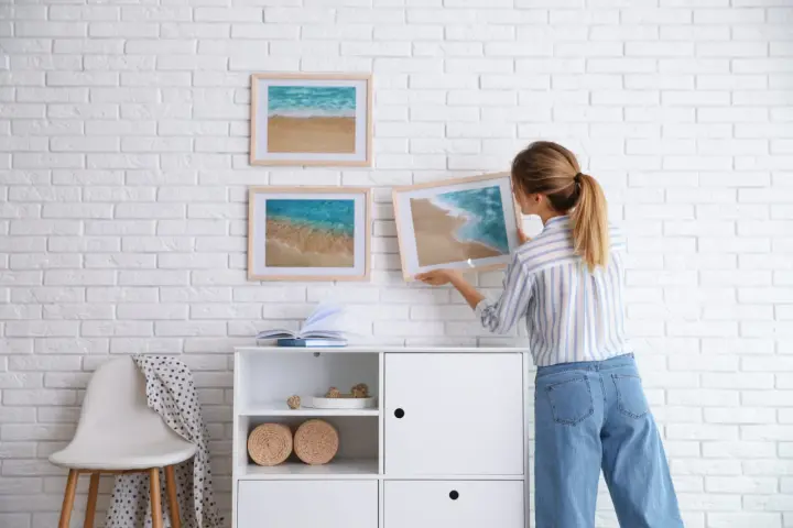 Mujer cuelga cuadros de paisajes marinos en pared de ladrillo blanco. Aprende como colocar cuadros de diferentes tamaños para un diseño armonioso.