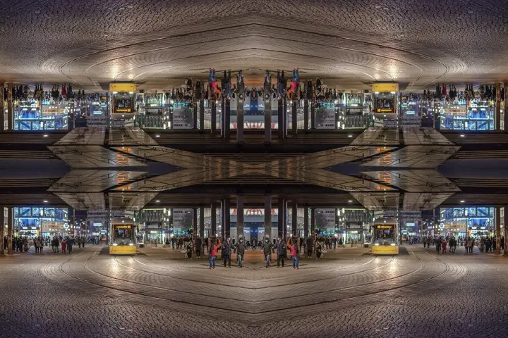 Nocne miasto z żółtym tramwajem i ludźmi. Kompozycja symetryczna tworzy surrealistyczny, odbity świat.