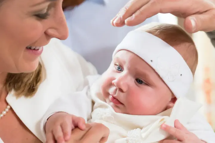 Uśmiechnięta mama trzyma dziecko w białej szatce. Dłoń ojca błogosławi malucha. Zastanawiasz się, ile kosztuje chrzest?