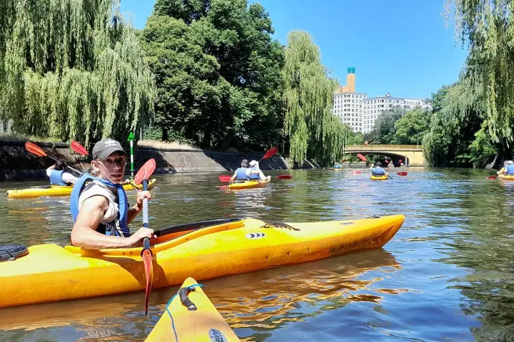 Eine Gruppe von Menschen paddelt auf gelben Kajaks auf einem Fluss in Berlin. Bäume säumen das Ufer, im Hintergrund ist ein Gebäude zu sehen.
