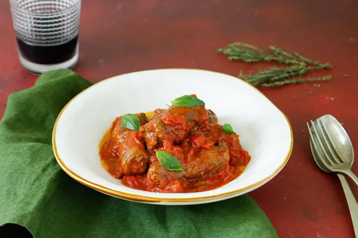 Involtini di carne in padella con sughetto, guarniti con basilico fresco, serviti in un piatto bianco su un tovagliolo verde.