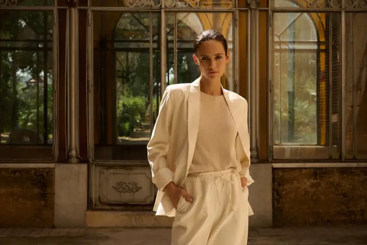 Mujer con blazer beige y pantalones de vestir, de pie frente a una estructura con ventanas.